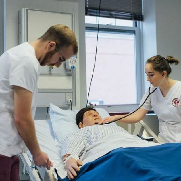 Photo of two nursing students standing over a medical bed holding a nursing simulation device
