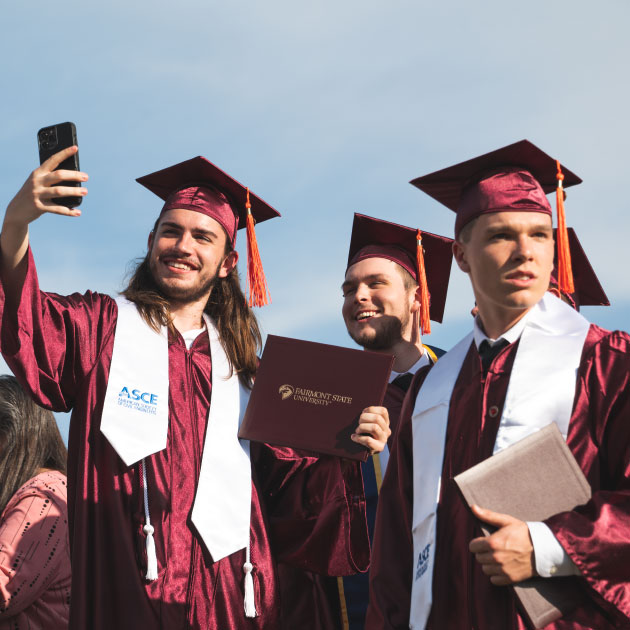Three graduates standing outdoors with a blue sky in the background, one holding up a cellphone to take a photo